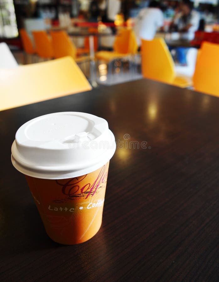 Ein Foto von einem Papier Tasse heißen Kaffee mit nehmen aus Kunststoff-Abdeckung angebracht, die auf das dunkle Holz Tisch in einem Uni-campus Kantine für Mitarbeiter und Studenten.