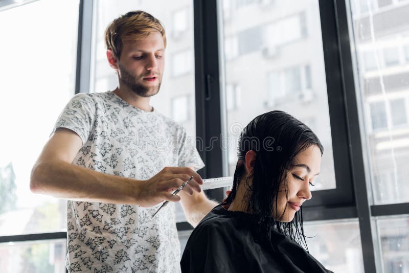 Coiffeur masculin de jeune hanche professionnelle coupant les cheveux foncés de la femme de client au salon image stock Coiffeur masculin de jeune hanche professionnelle coupant les cheveux foncés de la femme de client au salon image stock