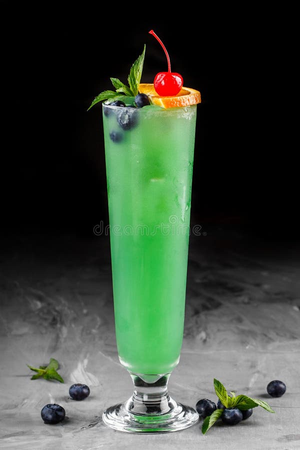 Cold green cocktail with blueberries, orange and ice in tall glass on dark background.