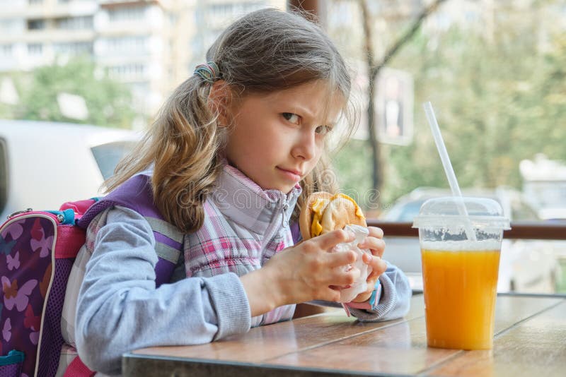 Colegiala bonita 8 del retrato al aire libre, 9 años con el cheeseburger y zumo de naranja, fondo del restaurante de los alimento imágenes de archivo libres de regalías Colegiala bonita 8 del retrato al aire libre, 9 años con el cheeseburger y zumo de naranja, fondo del restaurante de los alimento imágenes de archivo libres de regalías