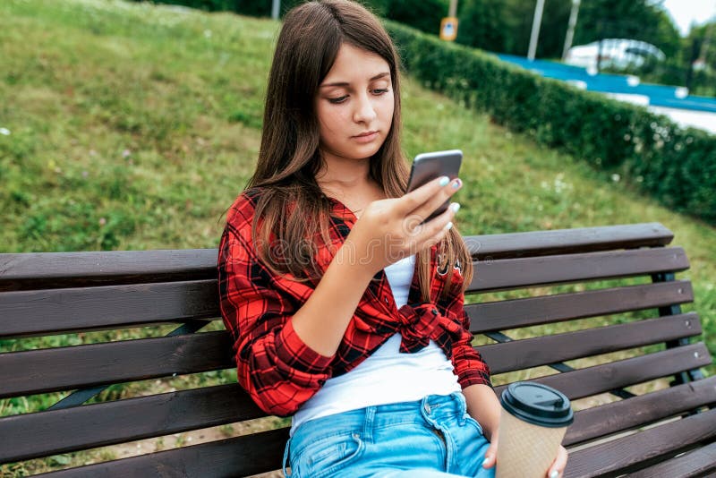 Colegiala de la muchacha 13-16 años que se sientan en un banco en su teléfono de la mano Uso en línea de la charla en las redes s fotos de archivo Colegiala de la muchacha 13-16 años que se sientan en un banco en su teléfono de la mano Uso en línea de la charla en las redes s fotos de archivo