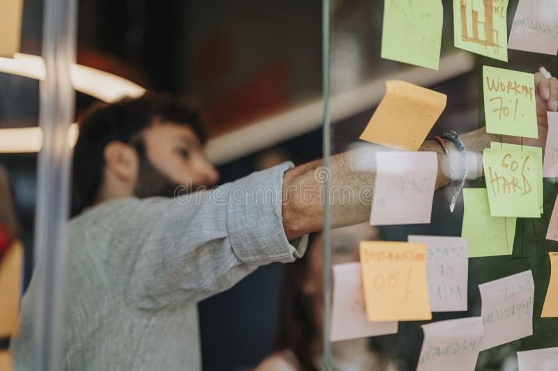 Collaboration en milieu de travail : partage de connaissances et d'idées d'affaires sur mur de verre avec des notes collantes. Un groupe diversifié d'hommes d'affaires brainstorming d'idées et partage des connaissances dans un bureau collaboratif. des notes collantes avec calculs et coûts de projet sont placées sur une paroi de verre.