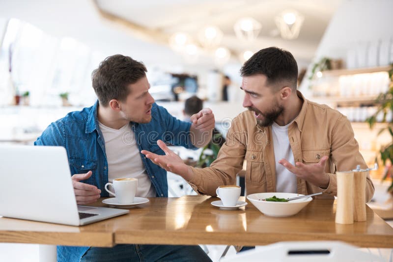 Colleagues men arguing about work issues during lunch break at cafe, one eats salad, another drinks coffee, working on project in