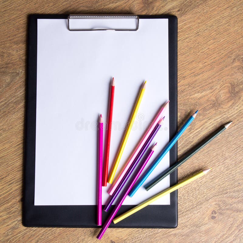 Colored drawing pencils and clipboard with blank paper on wooden
