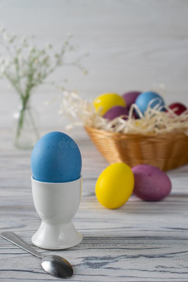 Colorful dyed easter eggs in a basket with blue egg in a cup in front on white wooden background
