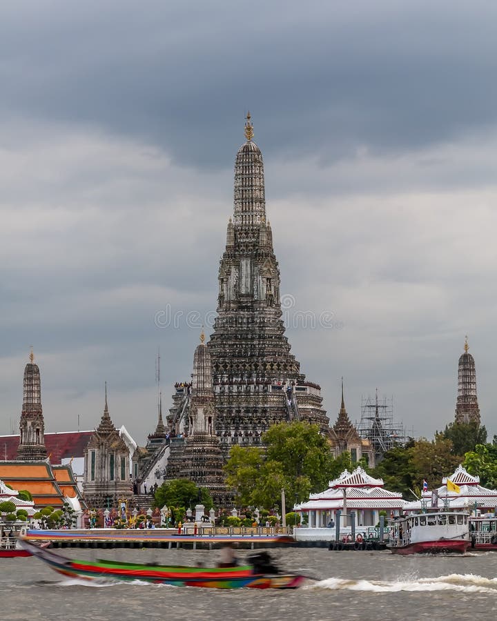 A colorful long tail boat sails on the Chao Phraya river in front of the Wat Arun temple, against cloudy sky, Bangkok, Thailand