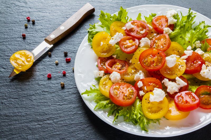 Colorful salad, fresh green leaves and sliced red and yellow cherry tomatoes, white plate, knife, black stone background