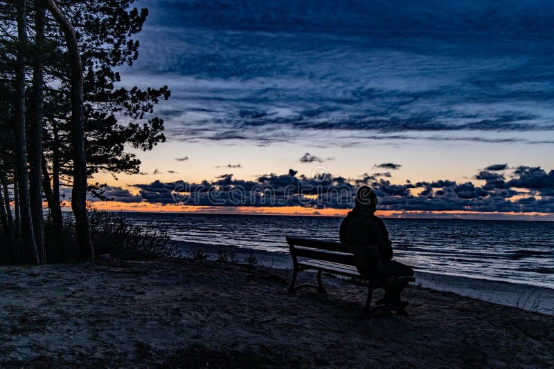 Colorful sunset on the sea observed by a man in a cap sitting on a black bench on a dune