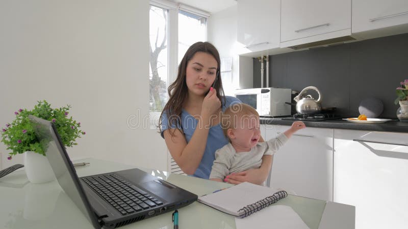 Combinant le travail et le parenting, mère avec pleurer peu de fils tout en travaillant sur l'ordinateur portable et parlant au t