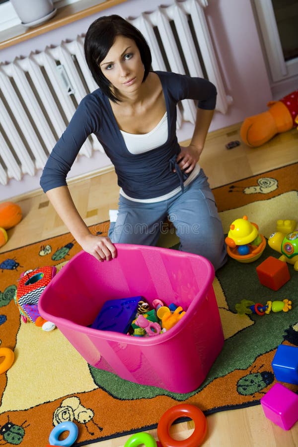 Young woman kneeling at container with toys and assembles strewn toys. High angle view. Young woman kneeling at container with toys and assembles strewn toys. High angle view.