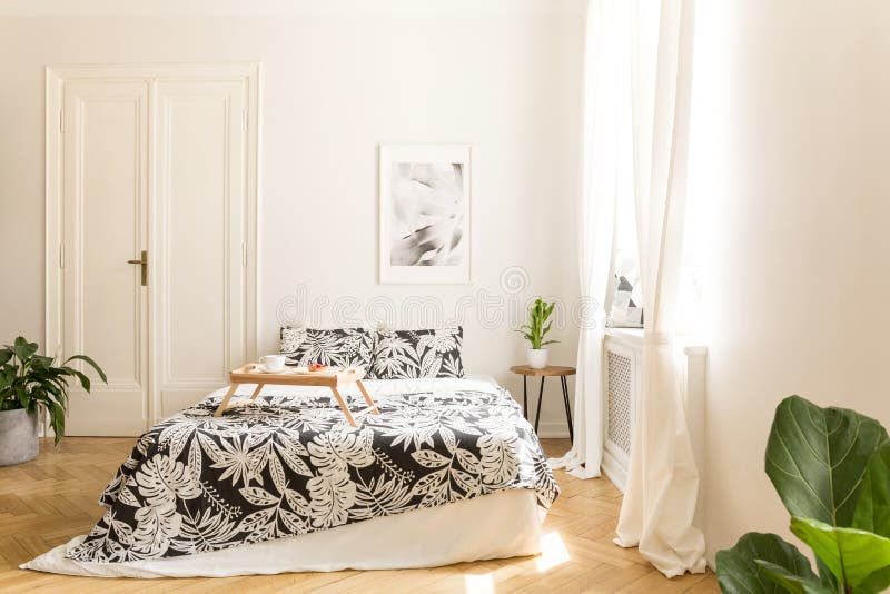 Comfortable big bed with white and black flower design bedding and a breakfast tray on in a wooden floor bright bedroom interior.