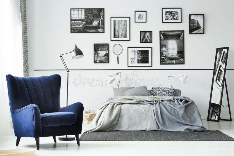 Comfortable blue armchair in monochromatic bedroom interior with king size bed and gallery of posters on the wall