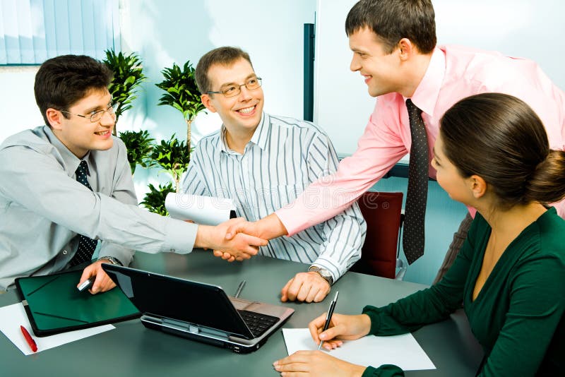 Photo of handshake of businesspeople with their smiling colleagues sitting near by in the office. Photo of handshake of businesspeople with their smiling colleagues sitting near by in the office