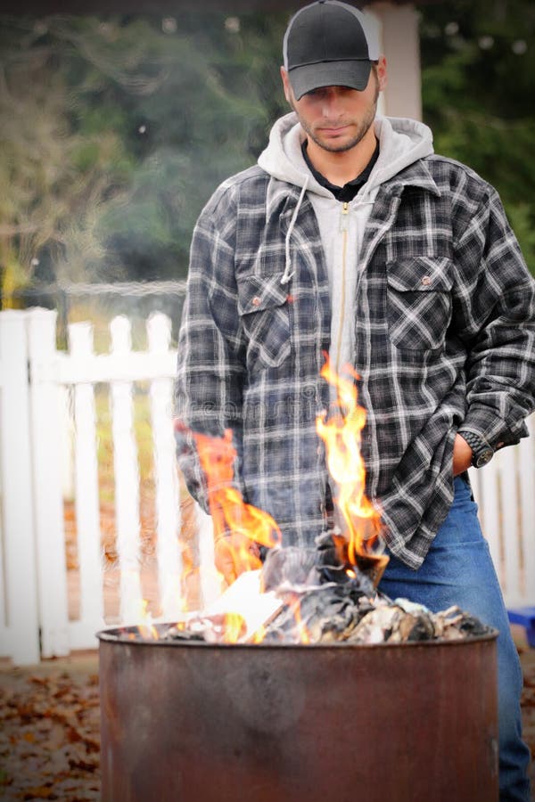 Man overseeing flames while burning garbage in an old fashioned steel burn barrel. Shallow depth of field with picket fence in background. Metal steel drum stock images, royalty-free photos and pictures