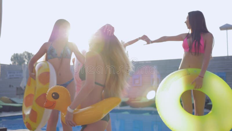 Company of slender long-haired girls in bathing suits dancing with inflatable rings near poolside at expensive resort on