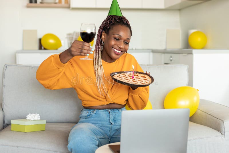 Compleanno online. donna di colore felice che festeggia il giorno mostrando la torta con candela e un bicchiere di vino alla webca fotografia stock libera da diritti