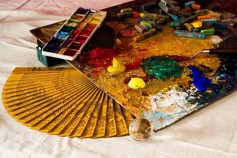 Composition of an artistic palette, hand fan, watercolors, acrylics, spatula, transparent ball and pastels