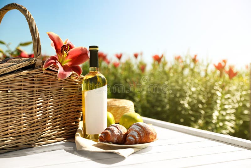 Composition with picnic basket and bottle of wine on wooden table in lily field. Space for text