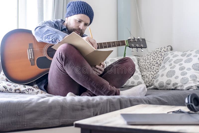 Compositor de sexo masculino entusiasta, anotando música en su cuaderno y sosteniendo una guitarra acústica, sentándose en un sof