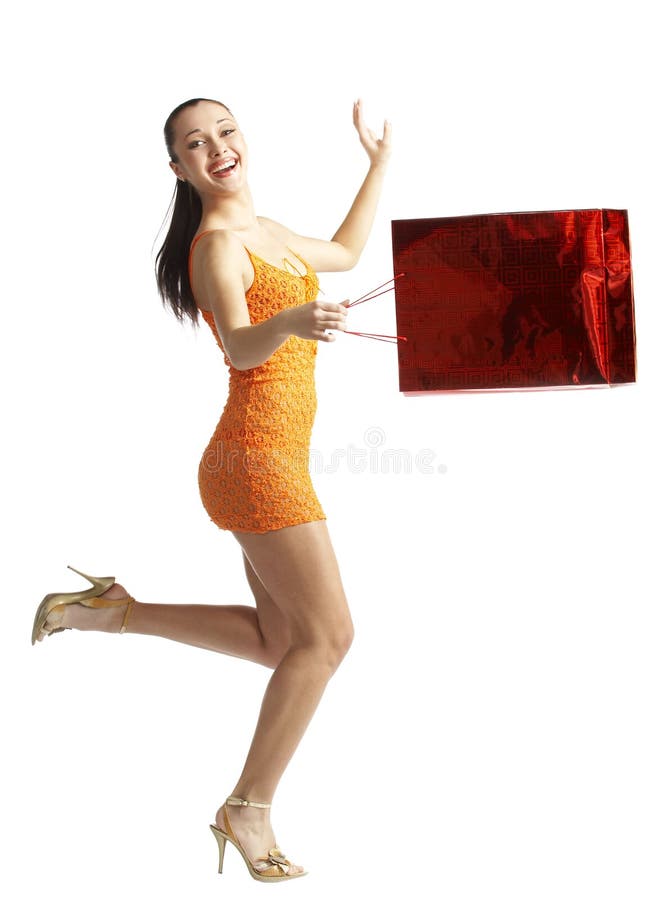 A young woman in an orange dress and gold heels, smiles widely as she swings her shopping bag while in mid-stride. A young woman in an orange dress and gold heels, smiles widely as she swings her shopping bag while in mid-stride.