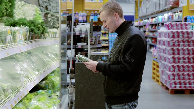 Comprando hierbas frescas en el supermercado. Un joven cliente con una chaqueta negra está leyendo la información en un perejil almacen de video