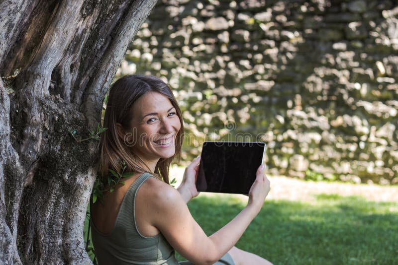 Compressa della lettura della donna e godere del resto in un parco sotto l'albero immagine stock libera da diritti Compressa della lettura della donna e godere del resto in un parco sotto l'albero immagine stock libera da diritti
