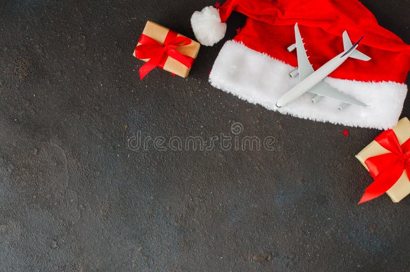 Trips or Christmas travel planning. Toy airplane on Santa hat and gift boxes on dark concrete. Top view or flat lay. Copy Space. Trips or Christmas travel planning. Toy airplane on Santa hat and gift boxes on dark concrete. Top view or flat lay. Copy Space.