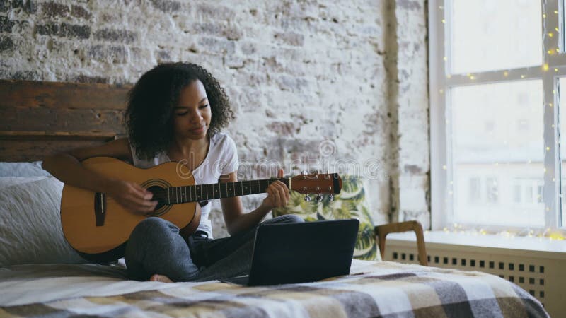 Concentraing Lernen des gelockten Afroamerikanerjugendlich-Mädchens, Gitarre unter Verwendung der Laptop-Computers zu spielen, di