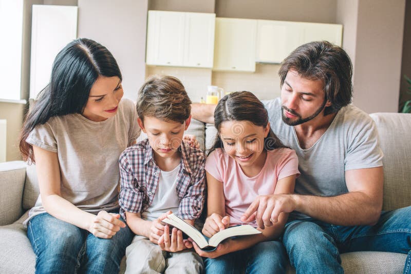 Concentrated kids are sitting on sofa with their parents and reading a book. Children hold it together. Guy is pointing on book