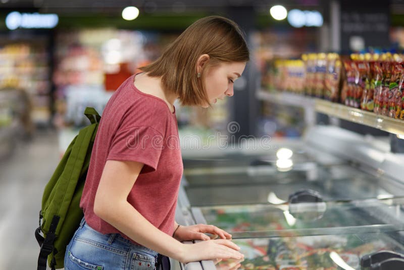 Concentrated lovely female teenager going to buy ice cream leans at fridge in supermarket, carries rucksack, dressed in casual clo stock photos Concentrated lovely female teenager going to buy ice cream leans at fridge in supermarket, carries rucksack, dressed in casual clo stock photos