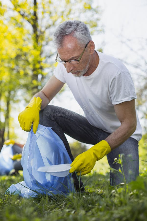 Concentrated mature volunteer finding eco project. Environmental task. Low angle of handsome mature volunteer wearing glasses and picking litter