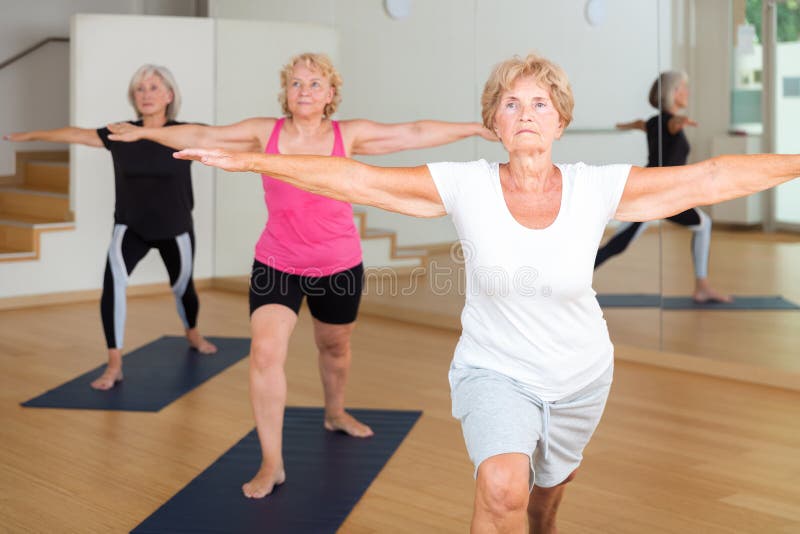 Concentrated senior woman performing standing lunging asana Virabhadrasana during yoga class with female group in fitness studio. Concentrated senior woman performing standing lunging asana Virabhadrasana during yoga class with female group in fitness studio.