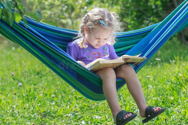 Concentrated two years old girl reading opened book on hanging hammock in green summer garden outdoors