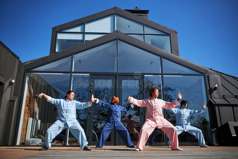 Group of people practicing tai chi in traditional Chinese clothes and doing exercises in front of a glass building on a terrace. Qigong class stock images, royalty-free photos and pictures