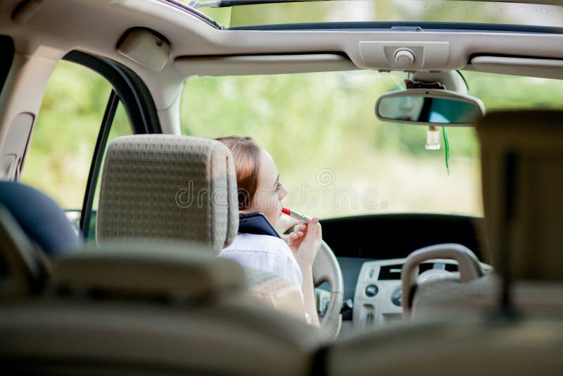 Concept of danger driving. Young woman driver red haired teenage girl painting her lips doing applying make up while driving the car.