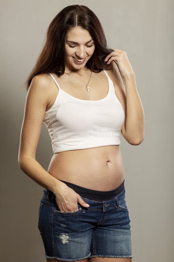 Concept de grossesse et d'attente - femme enceinte heureuse et contact de son ventre, fond gris, portrait image stock Concept de grossesse et d'attente - femme enceinte heureuse et contact de son ventre, fond gris, portrait image stock
