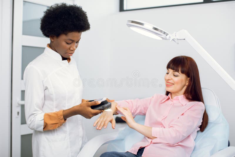 Skin cancer and melanoma prevention concept. Young confident African women doctor checking arm and body skin of young smiling pretty Caucasian women with dermatoscope in modern clinic. Skin cancer and melanoma prevention concept. Young confident African women doctor checking arm and body skin of young smiling pretty Caucasian women with dermatoscope in modern clinic