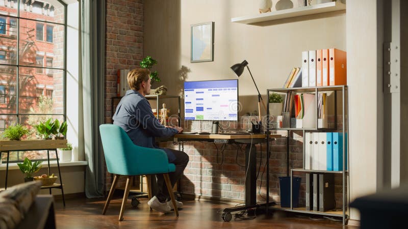 Remote Work Concept: Man Using Desktop Computer With Office Software to Check Schedule in Calendar. Young Male Planning Business Meetings in Stylish Loft Apartment with Big Window. Remote Work Concept: Man Using Desktop Computer With Office Software to Check Schedule in Calendar. Young Male Planning Business Meetings in Stylish Loft Apartment with Big Window.