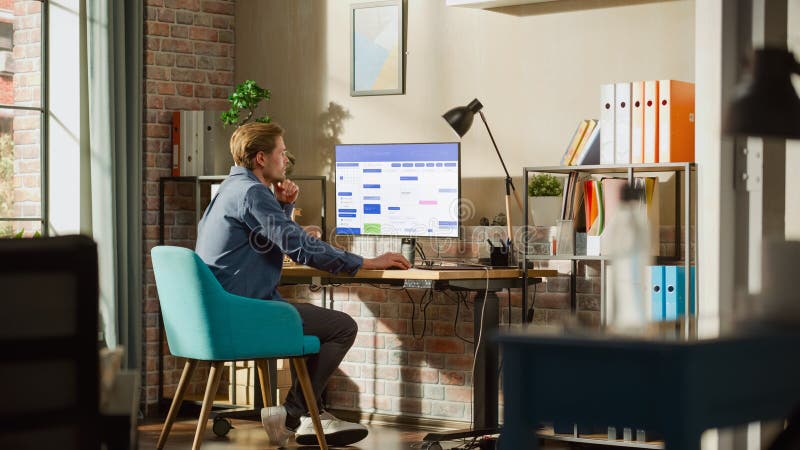 Remote Work Concept: Man Using Desktop Computer With Office Software to Check Schedule in Calendar. Young Male Planning Business Meetings in Stylish Loft Apartment with Big Window. Remote Work Concept: Man Using Desktop Computer With Office Software to Check Schedule in Calendar. Young Male Planning Business Meetings in Stylish Loft Apartment with Big Window.