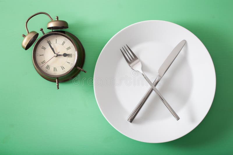 Concept of intermittent fasting, ketogenic diet, weight loss. fork and knife crossed on a plate and alarmclock. Concept of intermittent fasting, ketogenic diet, weight loss. fork and knife crossed on a plate and alarmclock