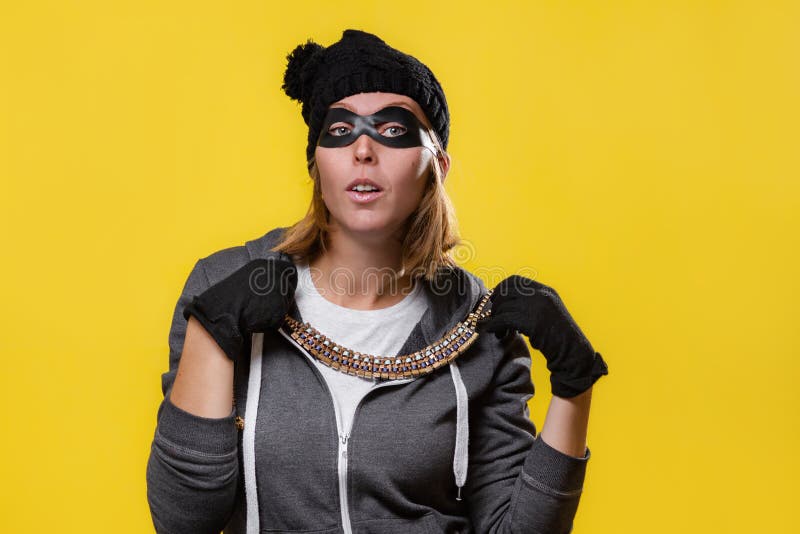 The concept of theft. A blonde woman in a hat, mask and gloves holds jewelry in both hands and tries on. Yellow background and