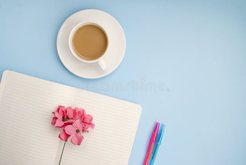 Concept workplace. White open notebook for notes, ballpoint pen, white cup of coffee with milk, pink flower on a blue background