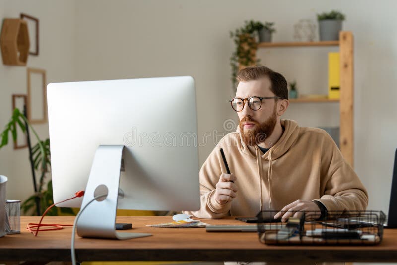 Concentrated young bearded designer in eyeglasses and hoodie sitting at desk and analyzing sketches on computer. Concentrated young bearded designer in eyeglasses and hoodie sitting at desk and analyzing sketches on computer