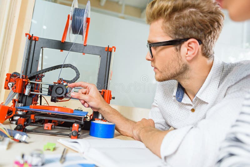 Young man is interested in 3d printer work. He is sitting at table and touching technology with curiosity. Young man is interested in 3d printer work. He is sitting at table and touching technology with curiosity