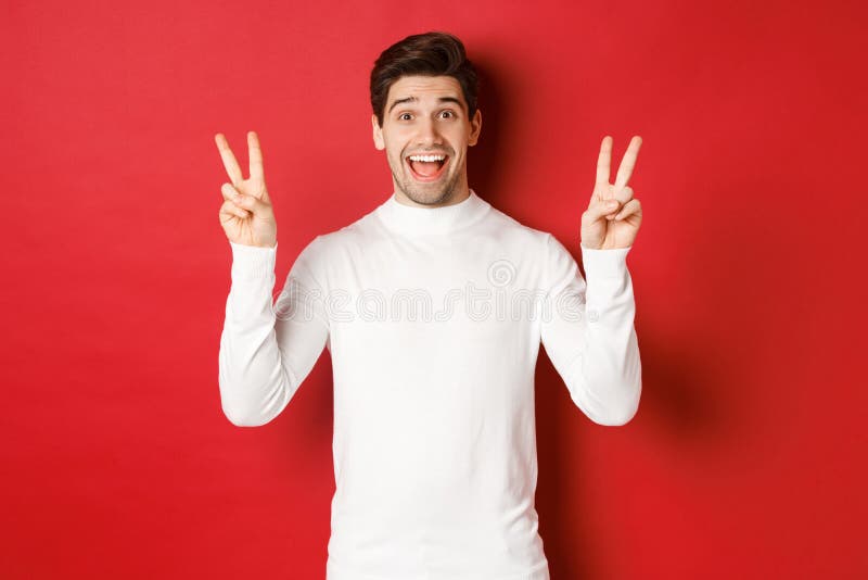 Concepto de navidad de invierno navidad navidad y estilo de vida. un tipo gracioso y guapo con suéter blanco mostrando signos de p. lindo tipo gracioso con suéter blanco mostrando signos de paz y sonriente alegre parado contra fondo rojo.