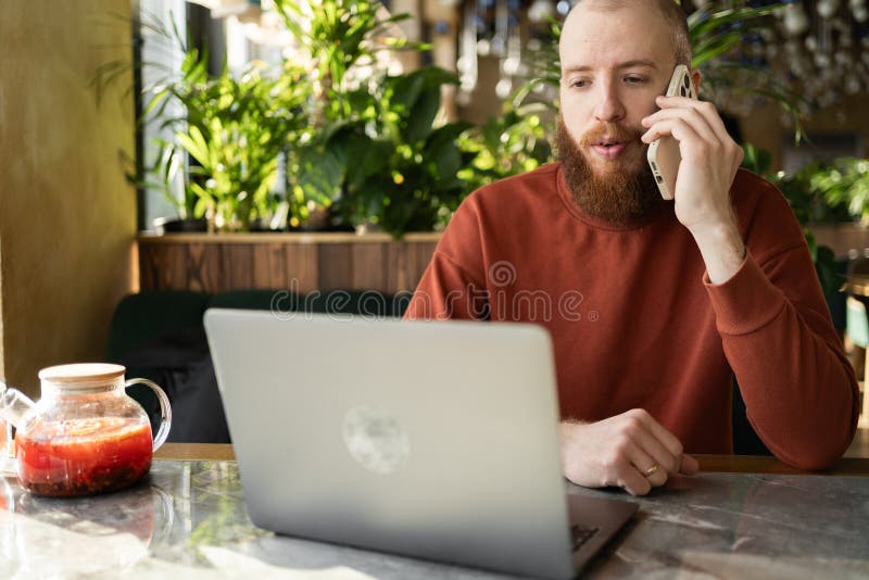 Concepto de trabajo remoto. Hombre con barba usando computadora portátil y teléfono móvil en conversación y comunicación. Freelanc imagen de archivo libre de regalías