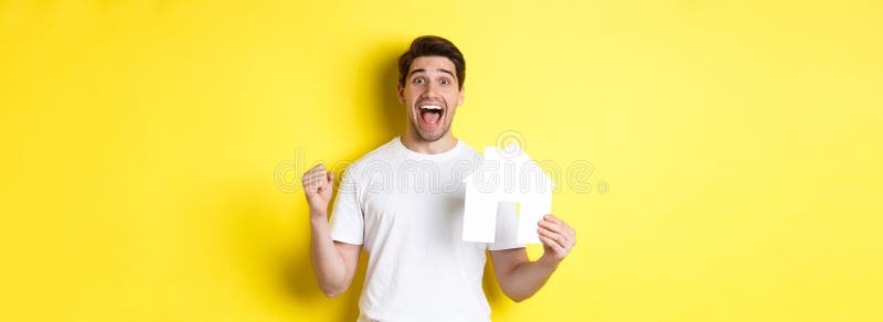 Concepto inmobiliario. hombre emocionado sosteniendo un modelo de casa de papel y celebrando estar feliz sobre fondo amarillo foto de archivo libre de regalías