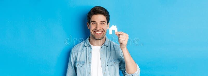 Concepto inmobiliario. joven y guapo hombre sonriendo mostrando una pequeña casa de papel burlándose buscando un departamento de p fotos de archivo libres de regalías