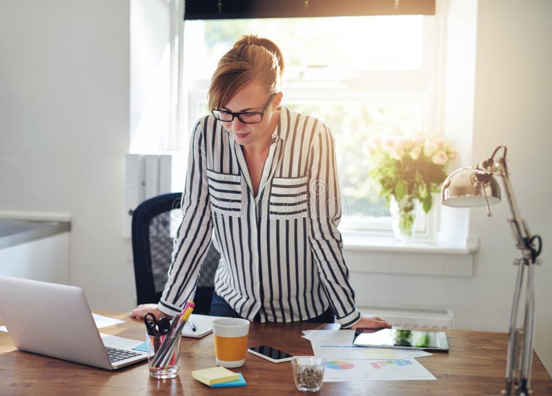 Concerned businesswoman studying information on the screen of her laptop computer, as she stands leaning on her desk in the office, in front of a bright window. Concerned businesswoman studying information on the screen of her laptop computer, as she stands leaning on her desk in the office, in front of a bright window