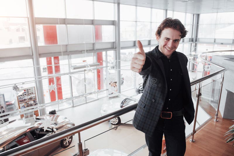 Concessionário automóvel de visita Homem novo no fundo do salão de beleza do carro O homem feliz mostra o dedo acima Venda do car imagem de stock Concessionário automóvel de visita Homem novo no fundo do salão de beleza do carro O homem feliz mostra o dedo acima Venda do car imagem de stock